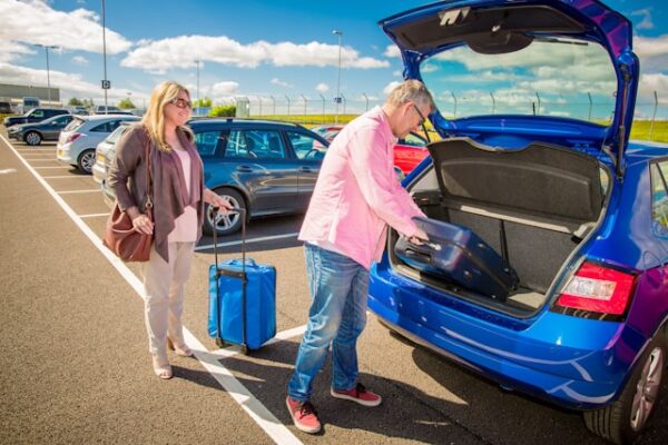 Muškarac stavlja kofer u gepek plavog auta dok žena drži kofer na parkingu